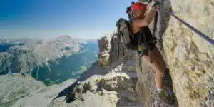 Mujer equipada con arnés y casco asegurada a un cable en una vía ferrata de alta montaña con vistas panorámicas.